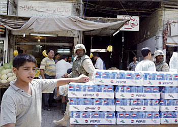 F_TO GO WITH AFP STORY: IRAQ-US-COMPANY-PEPSI A US soldier walks behind an Iraqi boy selling the US soft drink Pepsi along a street in Baghdad 20 June 2003. The Pepsi sold in Iraq is manufactured in Iraq under license. With Saddam Hussein's regime gone, an influx of foreign-made cola brands has poured into Baghdad, setting off a cut-throat soft drinks war against the dominant Iraqi Pepsi Cola. AFP PHOTO/Marwan NAAMANI