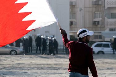 Sanabis, -, BAHRAIN : A Bahraini protestor waves his national flag as riot police stand guard (background) in the village of Sanabis near Manama on February 14, 2011 during a demonstration