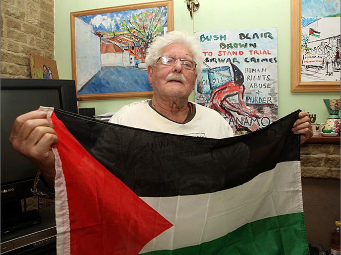 الناشط السياسي جون كات يرتدي قميص ضد بريطانيا وامريكيا ويحمل لعلم الفلسطيني وتظهر لوحات خلفه رسمها عن غونتاناموا وفلسطين