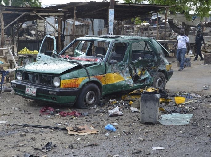 People walk past a damaged car at the site of a bomb explosion in Maiduguri, Nigeria, Friday, July 31, 2015. A woman suicide bomber killed many people at a crowded market early Friday in a blast that thundered across the northeastern Nigerian city of Maiduguri, witnesses said. (AP Photo/Jossy Ola)