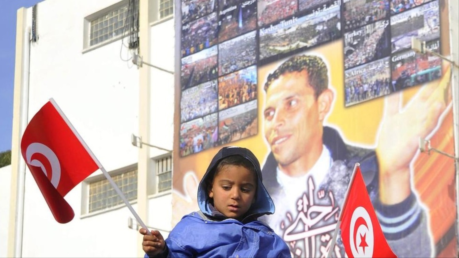 ‪طفل تونسي يحمل علم بلاده ووراءه صورة كبيرة لمحمد البوعزيزي‬ طفل تونسي يحمل علم بلاده ووراءه صورة كبيرة لمحمد البوعزيزي (الأوروبية- أرشيف)
