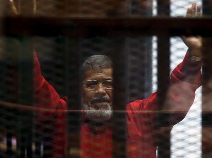 الرئيس المعزول محمد مرسي في البذلة الحمراء، الزي الخاص بالمحكومين بالإعدام، يُحيّي محاميه وأشخاص آخرين من وراء القضبان أثناء ظهوره في المحكمة في يونيو/حزيران 2015 مع أعضاء آخرين من الإخوان المسلمين، في ضواحي القاهرة.