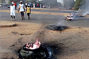 Guineans stroll by burning tires on the airport road in a suburb of Bissau 28 March 2004, the day of the legislative elections in Guinea Bissau. The elections to reconstitute parliament come six months after President Kumba Yala was deposed in a coup and a transitional government was put in place. Voting began late but calmly, and continued without incident, though the lack of voting materials in certain districts drew reactions of anger from frustrated would-be voters.