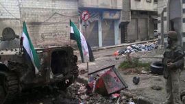 A picture made available on February 2, 2012 by the Syrian opposition Local Coordination Committees (LCC), is said to show a Syrian rebel standing next to a destroyed government forces tank bearing the rebel-adopted Syrian revolution flags, in the flashpoint city of Homs