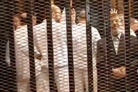 epa03935671 A grab made of video released by the Egyptian interior ministry showing ousted president Mohamed Morsi (R) standing behind bars with other defendants during his first trial session, in Cairo, Egypt, 04 November 2013. Egypt's toppled Islamist president, Mohammed Morsi, arrived on 04 November at the venue where his trial on charges of inciting the killing of opposition protesters is due to open. The so-called 'four-finger salute' has come to symbolize the lives lost during the dispersal of the Rabaa al-Adawiya protest camp by the Egyptian army. EPA/INTERIOR MINISTRY HANDOUT BEST QUALITY AVAILABLE HANDOUT EDITORIAL USE ONLY/NO SALES