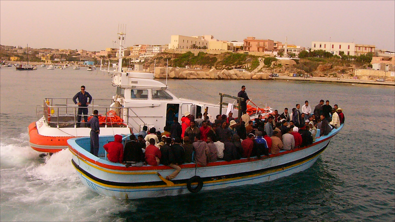 ‪قارب للاجئين عند دخوله جزيرة لامبيدوزا برفقة خفر السواحل الإيطالي (الجزيرة نت)‬ قارب للاجئين عند دخوله جزيرة لامبيدوزا برفقة خفر السواحل الإيطالي (الجزيرة نت)