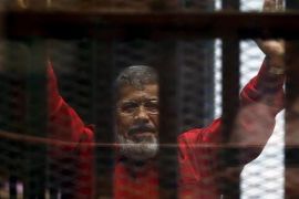 الرئيس المعزول محمد مرسي في البذلة الحمراء، الزي الخاص بالمحكومين بالإعدام، يُحيّي محاميه وأشخاص آخرين من وراء القضبان أثناء ظهوره في المحكمة في يونيو/حزيران 2015 مع أعضاء آخرين من الإخوان المسلمين، في ضواحي القاهرة.