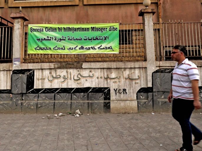 A picture taken on September 19, 2017 in the northeastern Syrian city of Qamishli shows a man walking past a banner reading in (top-bottom) Kurdish, Arabic, and Syriac: 'elections are the guarantee of the revolution of peoples', as Syrian Kurds prepare for the first local elections scheduled on September 22, 2017 in areas under their control within the framework of the federal system which was set up in March 2016. / AFP PHOTO / Delil souleiman (Photo credit sh