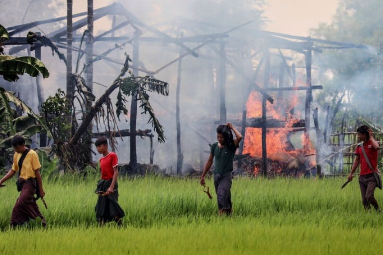 TOPSHOT - In this photograph taken on September 7, 2017, unidentified men carry knives and slingshots as they walk past a burning house in Gawdu Tharya village near Maungdaw in Rakhine state in northern Myanmar. The men were seen by journalists walking past the burning structure during a Myanmar government sponsored trip for media to the region. In the last two weeks alone 164,000 mostly Rohingya civilians have fled to Bangladesh, overwhelming refugee camps that were already bursting at the seams and scores more have died trying to flee the fighting in Myanmar's Rakhine state, where witnesses say entire villages have been burned since Rohingya militants launched a series of coordinated attacks on August 25, prompting a military-led crackdown. / AFP PHOTO / STR (Photo credit should read STR/AFP/Getty Images)