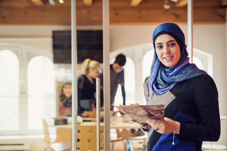 Muslim woman working in small fashion start-up enterprise. She is wearing an hijab, looking at the camera and smiling with papers in hands. Horizontal wait up indoors shot with copy space. This was taken in Montreal, Quebec, Canada.
