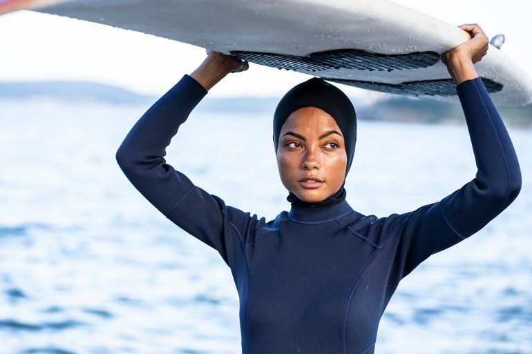Beautiful young woman in black wetsuit holding surfboard on beach.