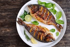 delicious roasted dorado or sea bream fish with lemon and orange slices, spices, fresh parsley and spinach on white platter on old dark wooden table view from above