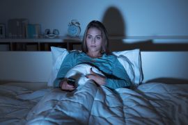 Young woman relaxing in bed late at night and watching tv, she is holding a remote control