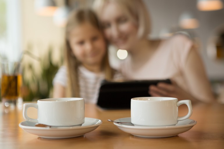 Two white cups of warm tea standing on table in cafe while mother and daughter sitting and watching video at background. Smiling woman and girl ordering cappuccino and coffee. Concept of lunch time.; Shutterstock ID 1419119429; purchase_order: aljazeera net; job: ; client: ; other: