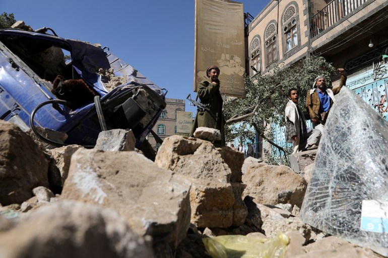Soldier and people stand by a vehicle destroyed by a Saudi-led air strike in Sanaa