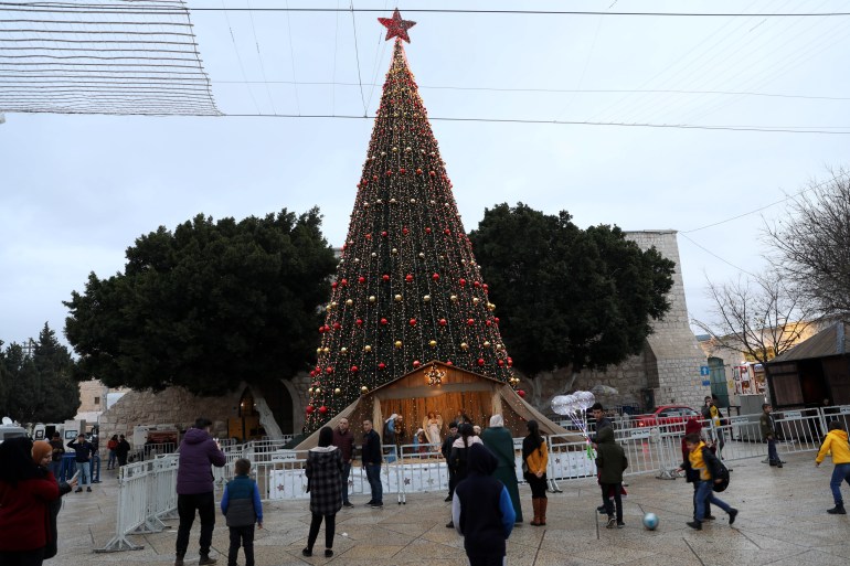 epa09655206 People look at the Christmas tree at the Manger Square next to the Church of Nativity, traditionally accepted as the birthplace of Jesus Christ, in the West Bank town of Bethlehem, 23 December 2021. The Church of the Nativity, built on the site where Jesus Christ is believed to have been born in the West Bank city of Bethlehem, is administered jointly by Greek Orthodox, Roman Catholic, Armenian Apostolic, and Syriac Orthodox church. EPA-EFE/ABED AL HASHLAMOUN
