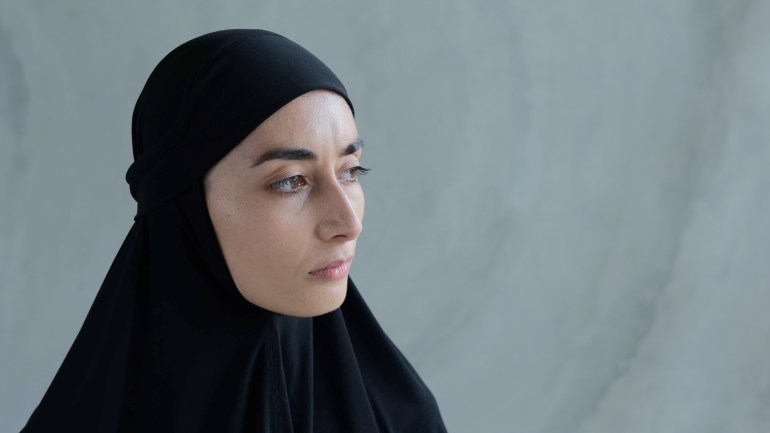 A Muslim woman with an expressive outline of the face, with black thick eyebrows, with beautiful brown eyes stands near a gray wall. The Arab woman wears a black hijab on her head. GettyImages-1390790892