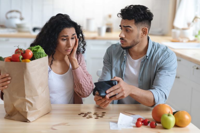 Coronacriris Concept. Depressed Arab Couple In Kitchen Counting Remaining Money After Food Shopping, Upset Middle Eastern Spouses Looking At Empty Wallet, Shocked By Big Prices For Grocery, Closeup shutterstock_1974885530