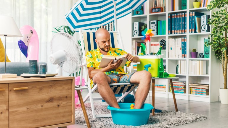 Man spending his summer vacations at home, he is sitting on a deckchair in the living room and reading a book GettyImages-1344258342