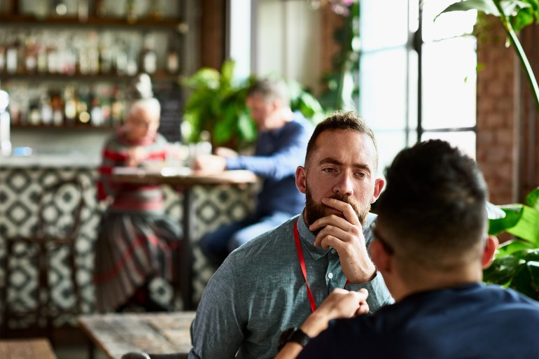 Man listening thoughtfully to business colleague in restaurant - stock photo Over shoulder view of mid adult man with beard listening to friend with hand on chin, contemplation, empathy, care GettyImages-1181221241