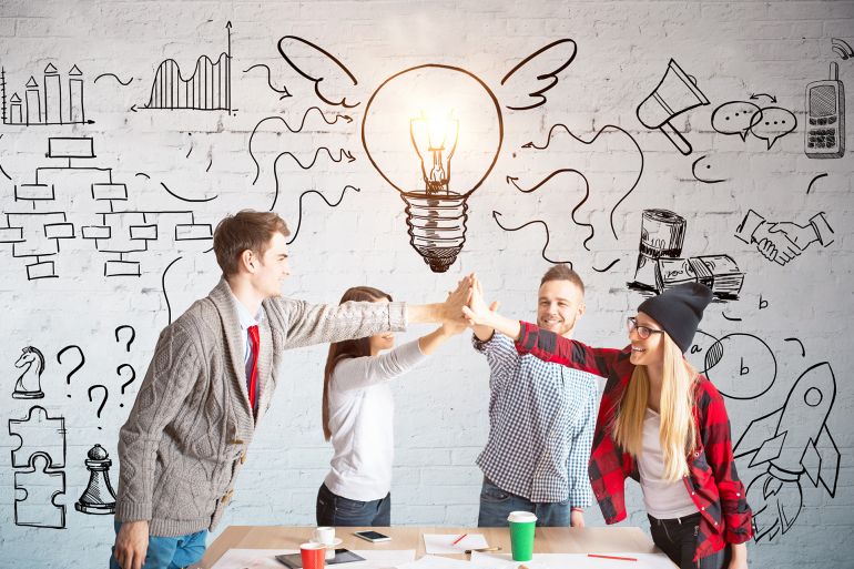 Young european team members hi-fiving each other above wooden desk. Brick wall with business sketch in the background. Teamwork concept shutterstock_658767439