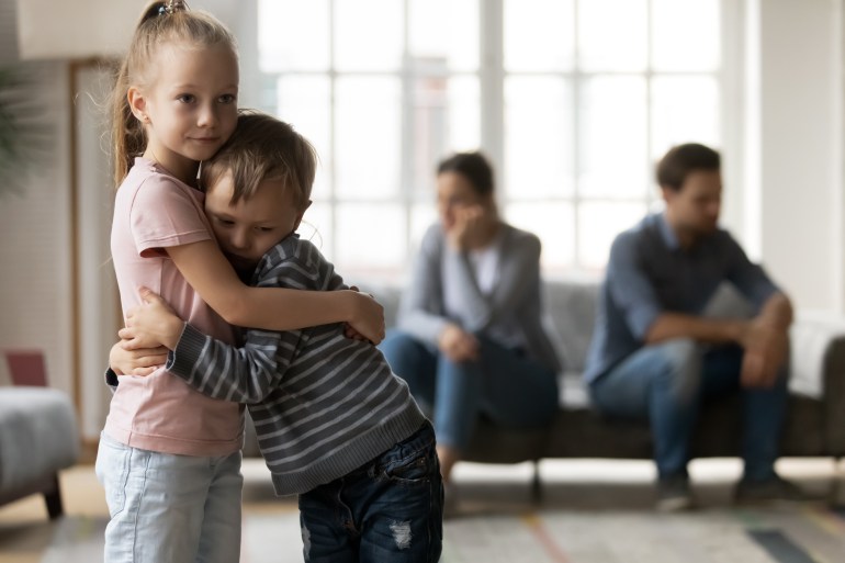 Upset little girl and boy, sister and brother hugging, suffering from parents quarrel close up, family conflict, offended mother and father ignoring each other after argument, children and divorce shutterstock_1755709433