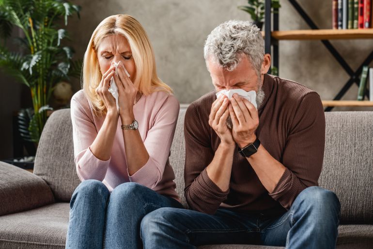 Middle aged couple sitting on sofa blowing their nose , flu concept with tissues and blanket
