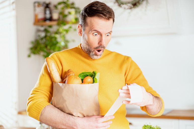 GettyImages-1369602921 Surprised man looking at store receipt after shopping, holding a paper bag with healthy food. Guy in the kitchen. Real people expression. Inflation concept.