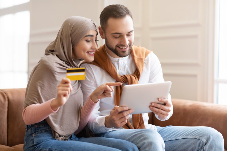 Happy Muslim Couple Shopping Online Using Tablet Computer And Credit Card Buying Things Sitting On Sofa At Home. Joyful Consumers, Sales And Modern Internet Shopping Concept. Selective Focus
