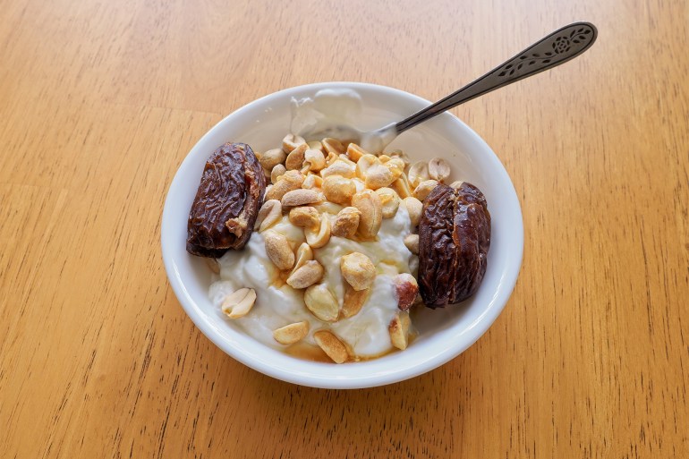A healthy decadent snack of Greek yogurt with peanuts and pitted dates drizzled with honey - stock photo A healthy decadent snack of Greek yogurt with peanuts and pitted dates drizzled with honey. The Deglet Nour dates are large and succulent in white bowl with spoon on hardwood table. GettyImages-1445274642