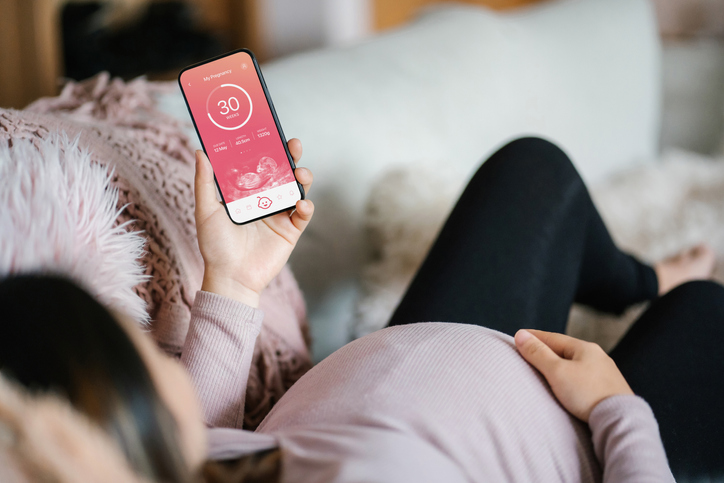 Young Asian pregnant woman lying on sofa in the living room at cozy home, using baby tracker app on smartphone to monitor her baby's growth and development throughout the pregnancy journey. Technology and pregnancy lifestyle