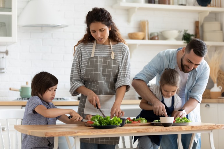 Caring father and mother teaching little children top use knife, standing at table in kitchen. Happy dad and mom with kids chopping vegetables lettuce on wooden cutting board, preparing dinner.