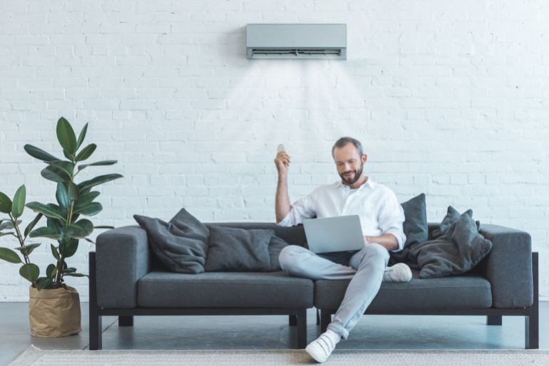 handsome man turning on air conditioner with remote control while using laptop on sofa at home; Shutterstock ID 1136979317; purchase_order:Creative Team ; job:; client:; other: