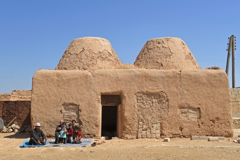 A family sits outside their traditional mud-brick house, known as 'beehive house', in the village of Umm Amuda al-Kabira in Aleppo's eastern countryside, north of Damascus on August 11, 2023. - Traditional mud houses that residents of northern Syria have built for thousands of years risk disappearing, as 12 years of war have emptied villages and left the homes crumbling. The village in Aleppo province is among a handful of villages where residents long used to live in small domed houses made of mud bricks and brittle hay. (Photo by AFP)