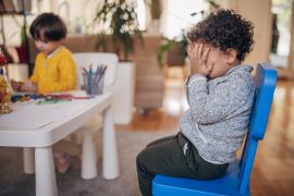 Two people, girl playing while boy is crying in preschool classroom.