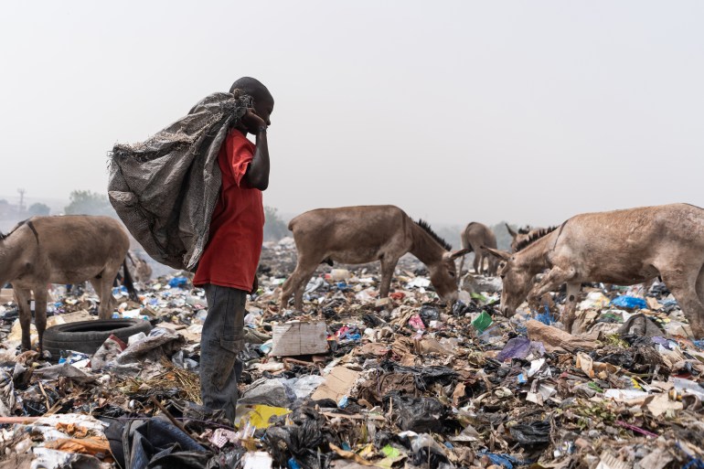 Lonely African boy standing in a landfill with a black plastic bag on his shoulder looking for reusable material, ...