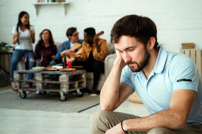 Young student guy feels upset and isolated while his friends celebrating party at home