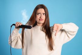 Upset young woman with flattening iron showing thumb down on light blue background. Hair damage