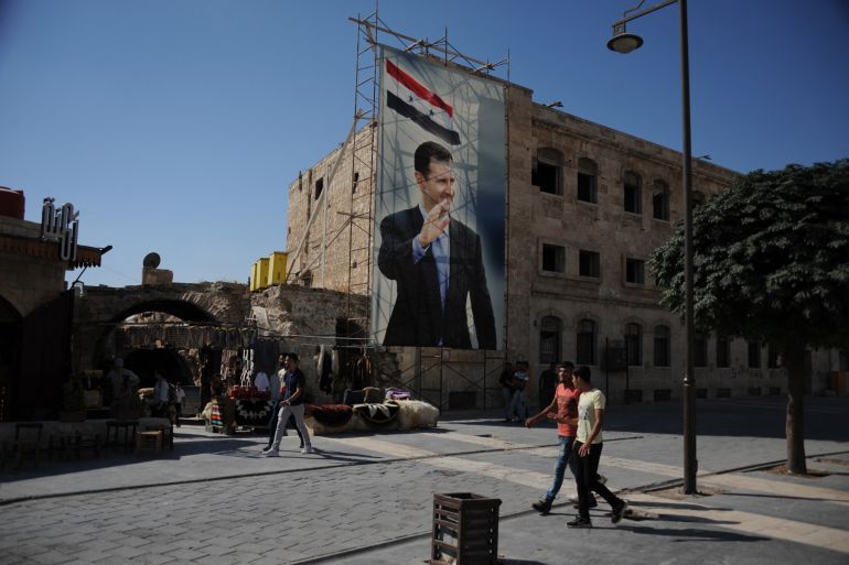 A picture taken during a guided tour with the Russian army shows men walking past a poster of Syrian President Bashar al-Assad in the old city of Aleppo on September 27, 2019. / AFP / Maxime POPOV