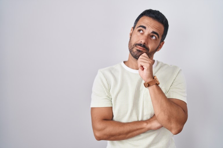 Hispanic man with beard standing over isolated background with hand on chin thinking about question, pensive expression. smiling with thoughtful face. doubt concept.