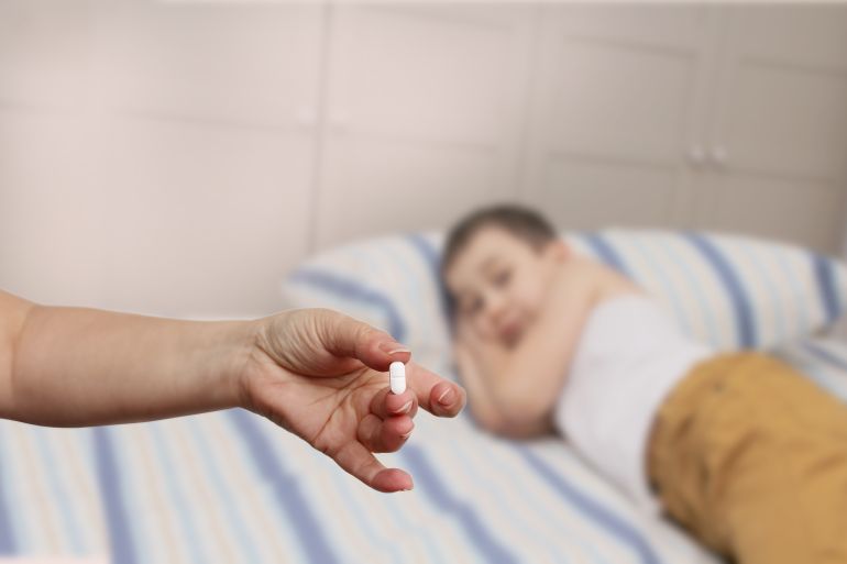 female hand holds out, offering, white elongated pill, close-up, blurred image of a lying child in the background, copy space, concept of medical care, treatment
