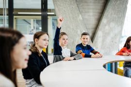A young boy raising his hand to ask a question while attending a tech workshop for kids with his classmates.