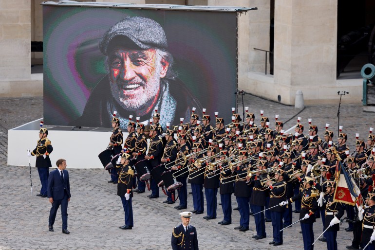 France's President Emmanuel Macron review troops as a photograph of late French actor Jean-Paul Belmondo appears on a giant screen during a national tribute ceremony at the Hotel des Invalides in Paris, on September 9, 2021. Several hundred of attendees have gathered to pay their respects to one of the most popular French actors : his relatives, the French President and members of the government as well as personalities from the world of cinema, culture and sport. Belmondo died on September 6, 2021 aged 88. (Photo by Ludovic MARIN / AFP)