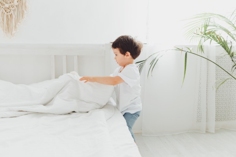 Happy little Asian boy cleans his room and folds a blanket in the bedroom at home, The kid makes the bed, adjusts the blanket on the big bed; Shutterstock ID 2227431237; purchase_order: aljazeera ; job: ; client: ; other: