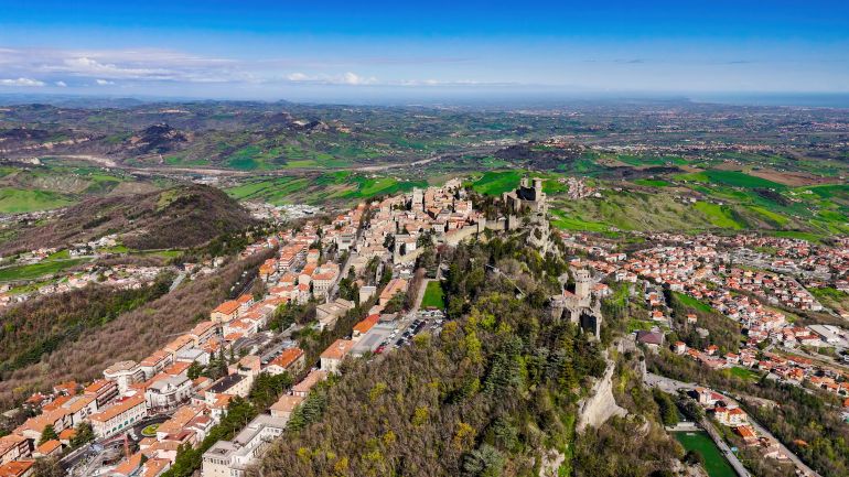 This aerial image captures a sweeping perspective of Guaita and Cesta Towers, the first and second medieval towers of San Marino, perfectly aligned along the rocky spine of Monte Titano. Guaita Tower, closest to the viewer, was originally built in the 11th century and functioned as both a fortress and prison, while Cesta Tower, further behind, stands on the highest peak and houses a museum of historical weaponry. The composition showcases the towers’ strategic elevation above the forested slopes and surrounding settlements, symbolizing San Marino’s enduring independence, historical resilience, and architectural heritage.