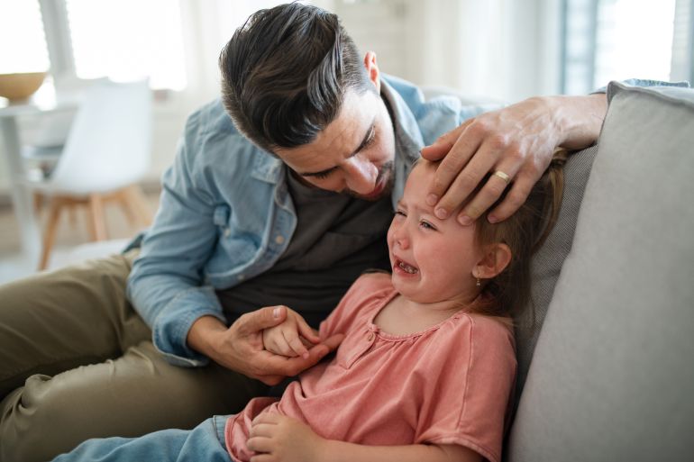 Young man sitting on sofa, holding and calming down his little daughter indoors at home.